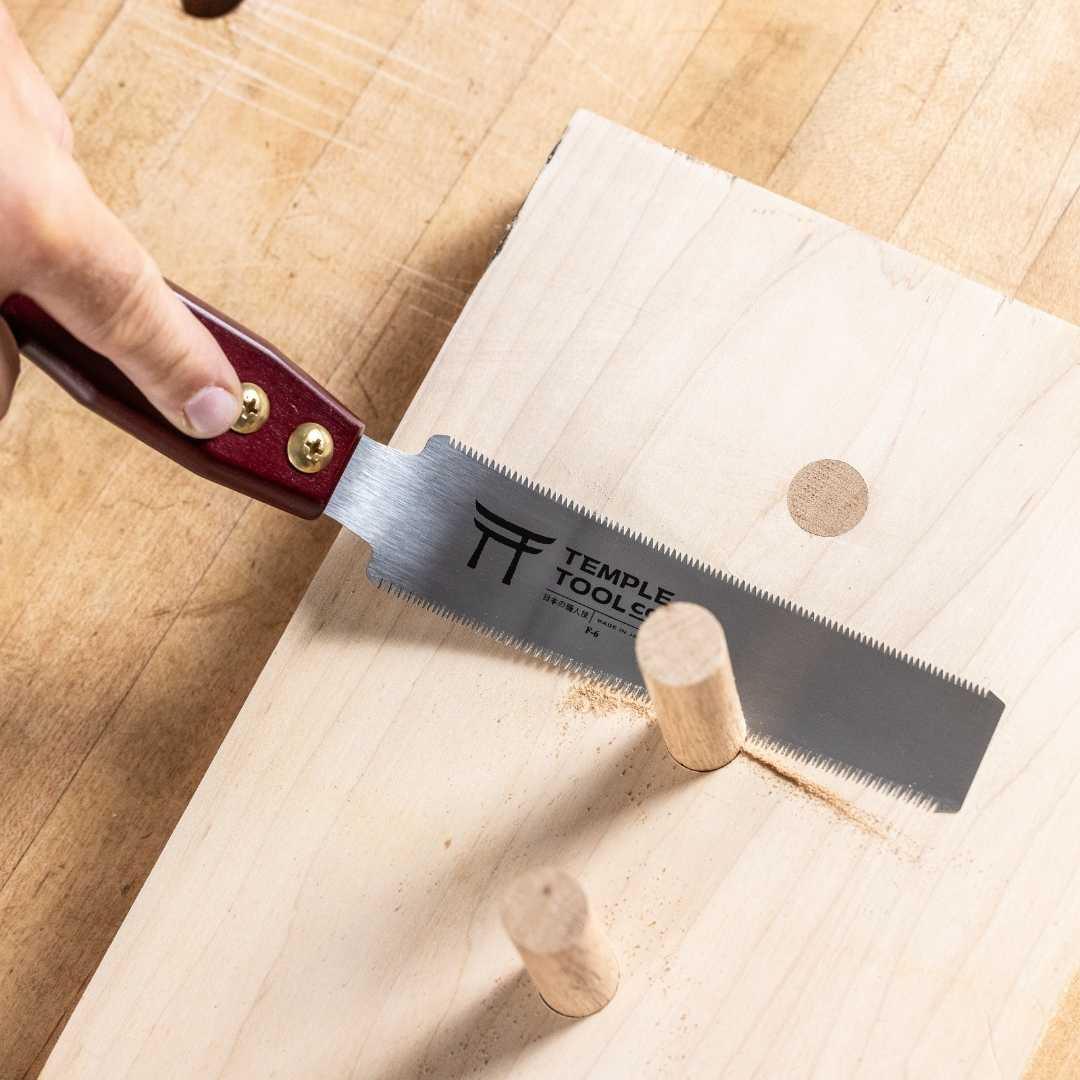 Hand using Temple Tool flush cut pull saw with red handle to trim wooden dowels on light wood board