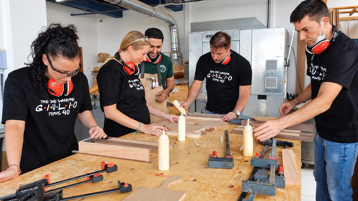 Team in matching t-shirts and ear defenders gluing hardwood boards together at a workbench while a Makers Society maker guides them
