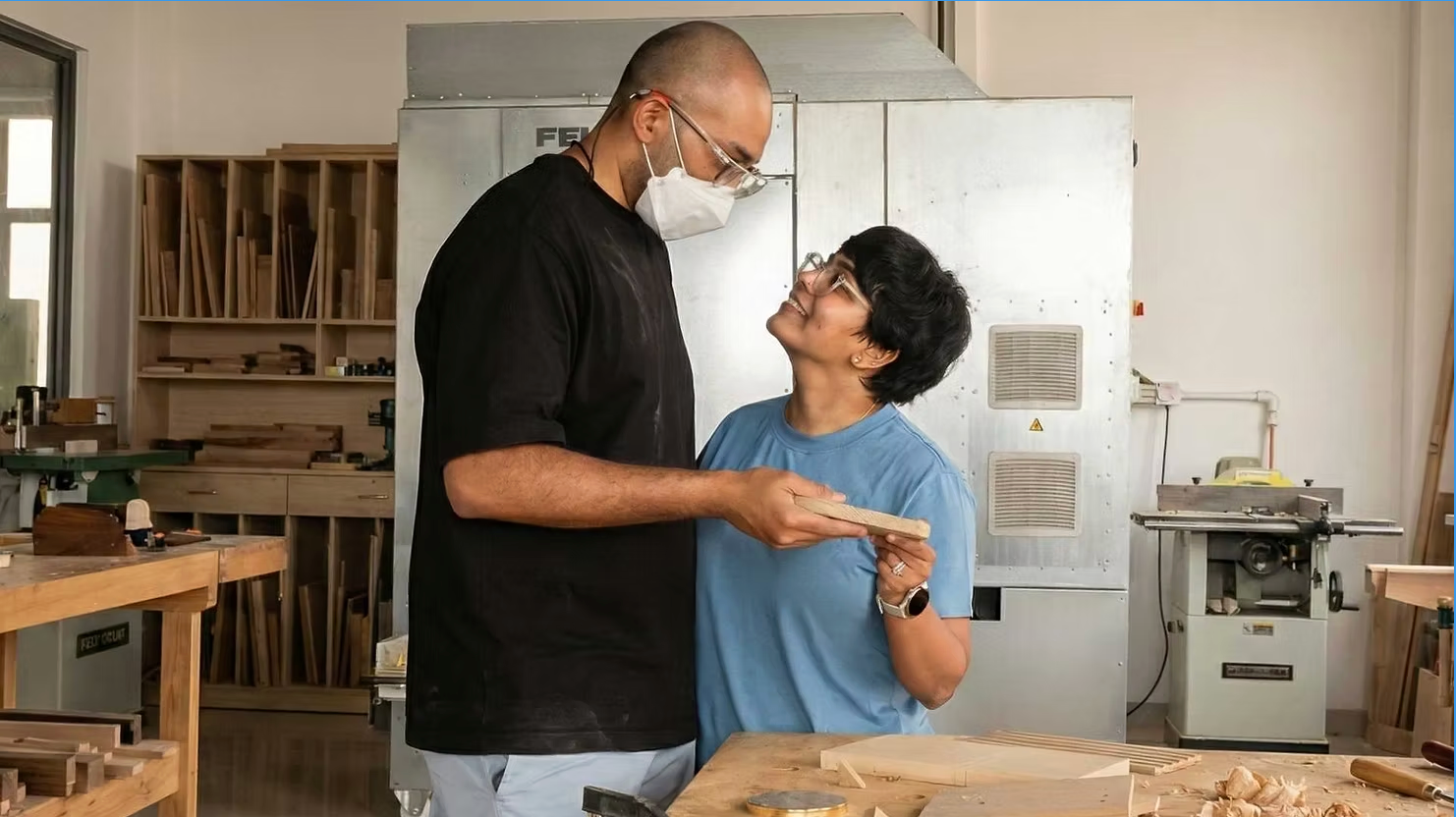 Couple in a woodworking shop holding a small wood piece and smiling at each other near a workbench