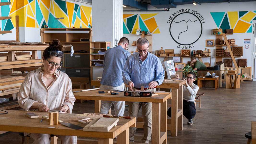 Adults at workbenches, woman chiselling in foreground, TMS logo visible