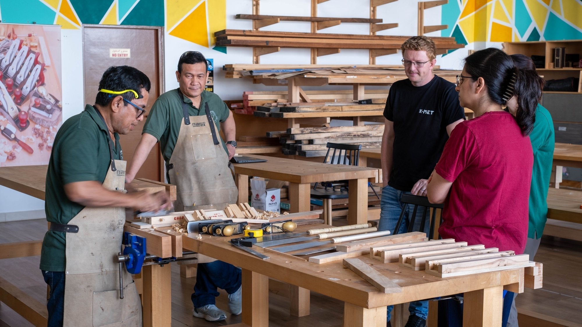 Group gathered around a workbench at the start of a woodworking workshop in Dubai, timber racks behind them