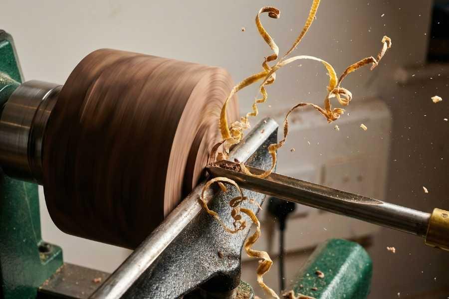 A close-up action shot of a woodturning lathe at The Makers Society, featuring a sharp gouge tool cutting into spinning dark walnut wood with long, curly golden shavings flying in mid-air against a workshop background.