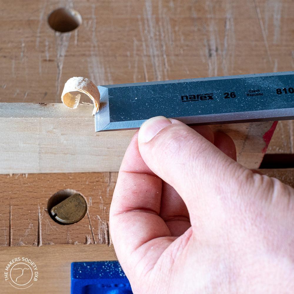 Hand holding a Narex wood chisel carving a wood shaving on a wooden workbench in a woodworking setting
