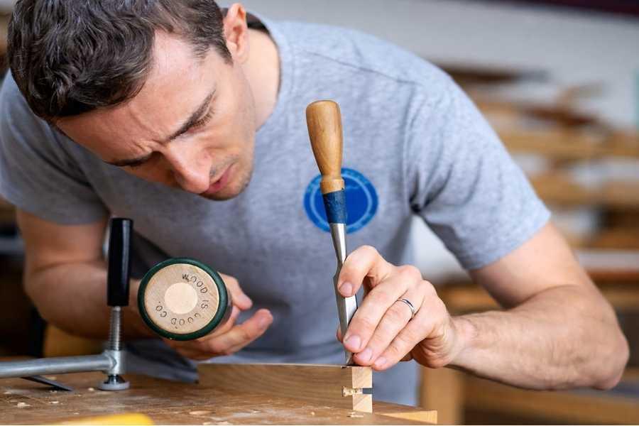 Man carefully chiseling a dovetail joint in a wooden board at a workbench, using a chisel and wooden mallet in a professional woodworking workshop.