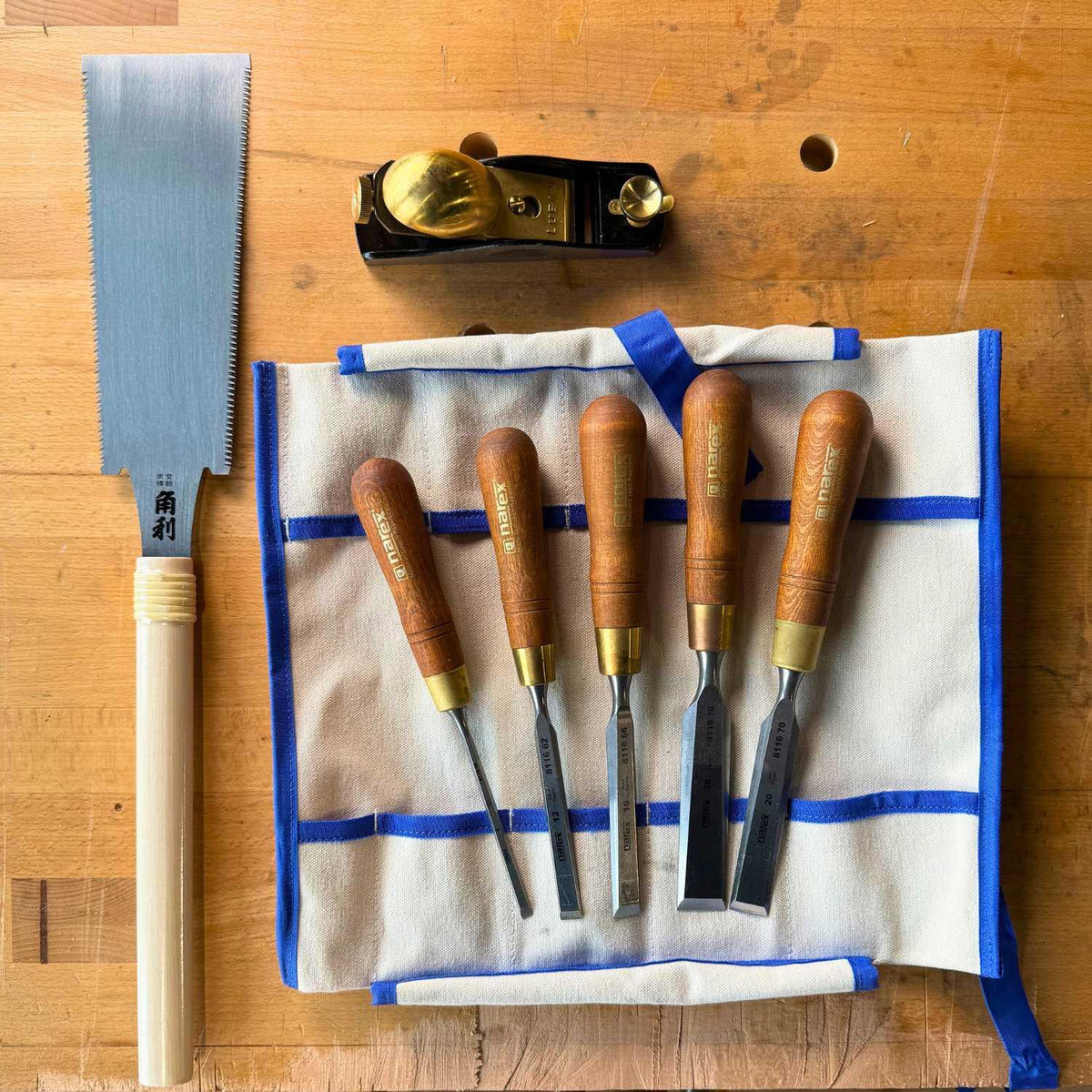 Woodworker's starter kit with Japanese saw, wooden-handled chisels on fabric roll, and brass hand plane on wooden workbench