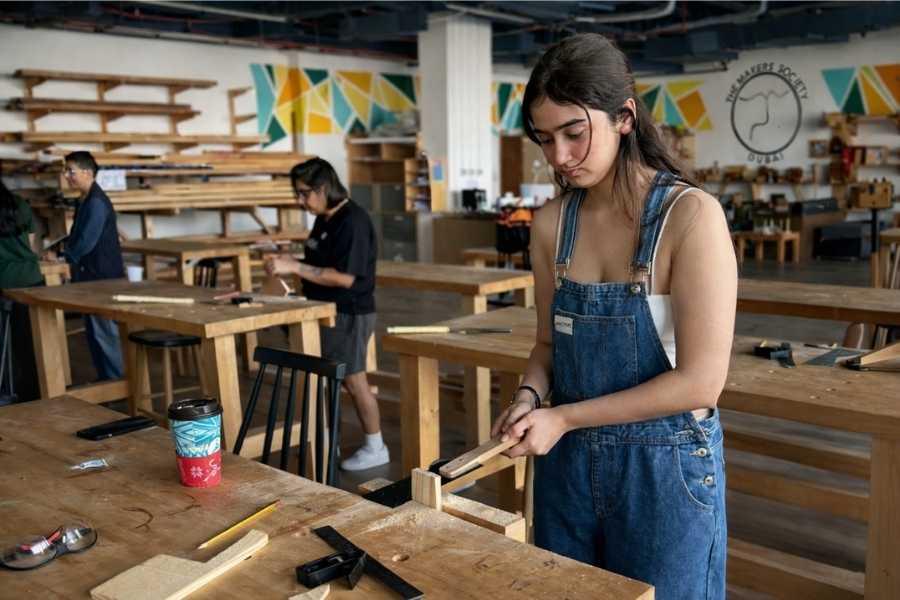 Young woman using a chisel to shape wood at a workbench inside a professional woodworking studio, with other participants working in the background at The Makers Society Dubai.