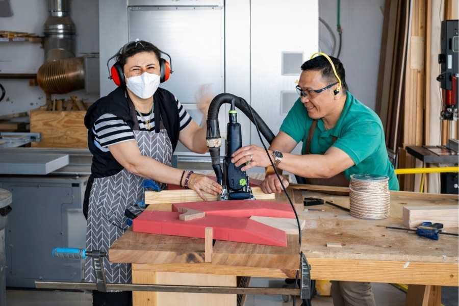 Two adults wearing safety gear collaboratively using a power router on a wooden workpiece at The Makers Society Dubai