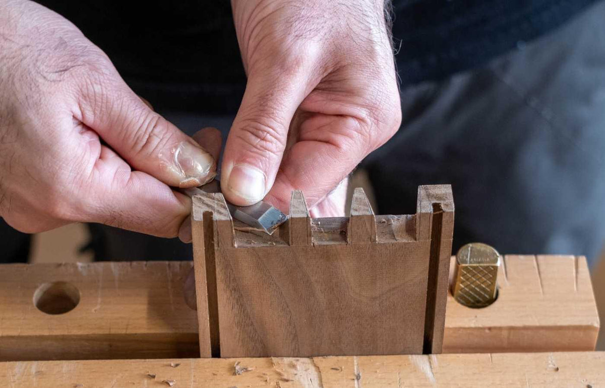 Close-up of hands using a dovetail chisel to carve precise joinery on walnut wood in a woodworking workshop
