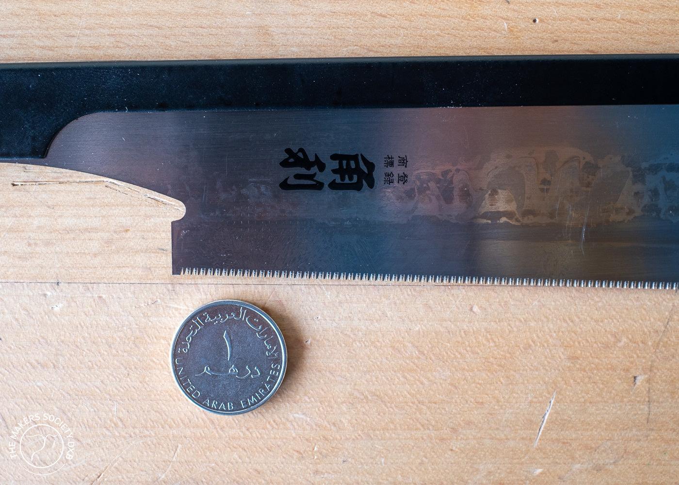 Close-up of a 210mm Kakuri Sakura handle Dozuki saw blade with fine teeth beside a UAE 1 dirham coin on a wooden surface