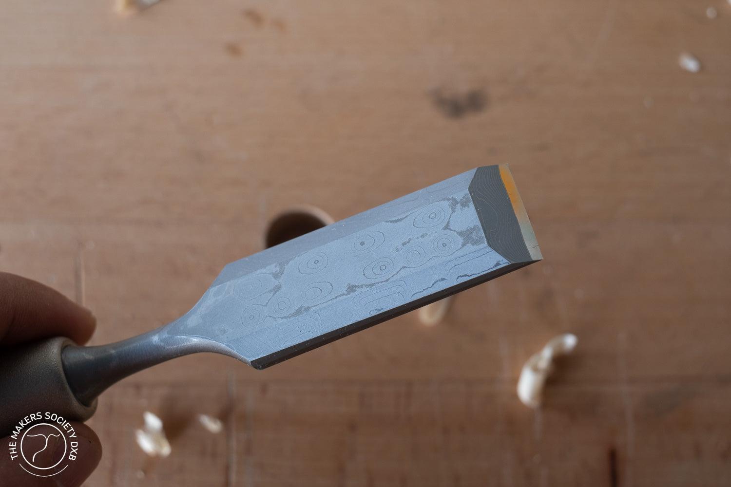 Hand holding a premium Damascus woodworking chisel with distinct patterned blade on wooden background