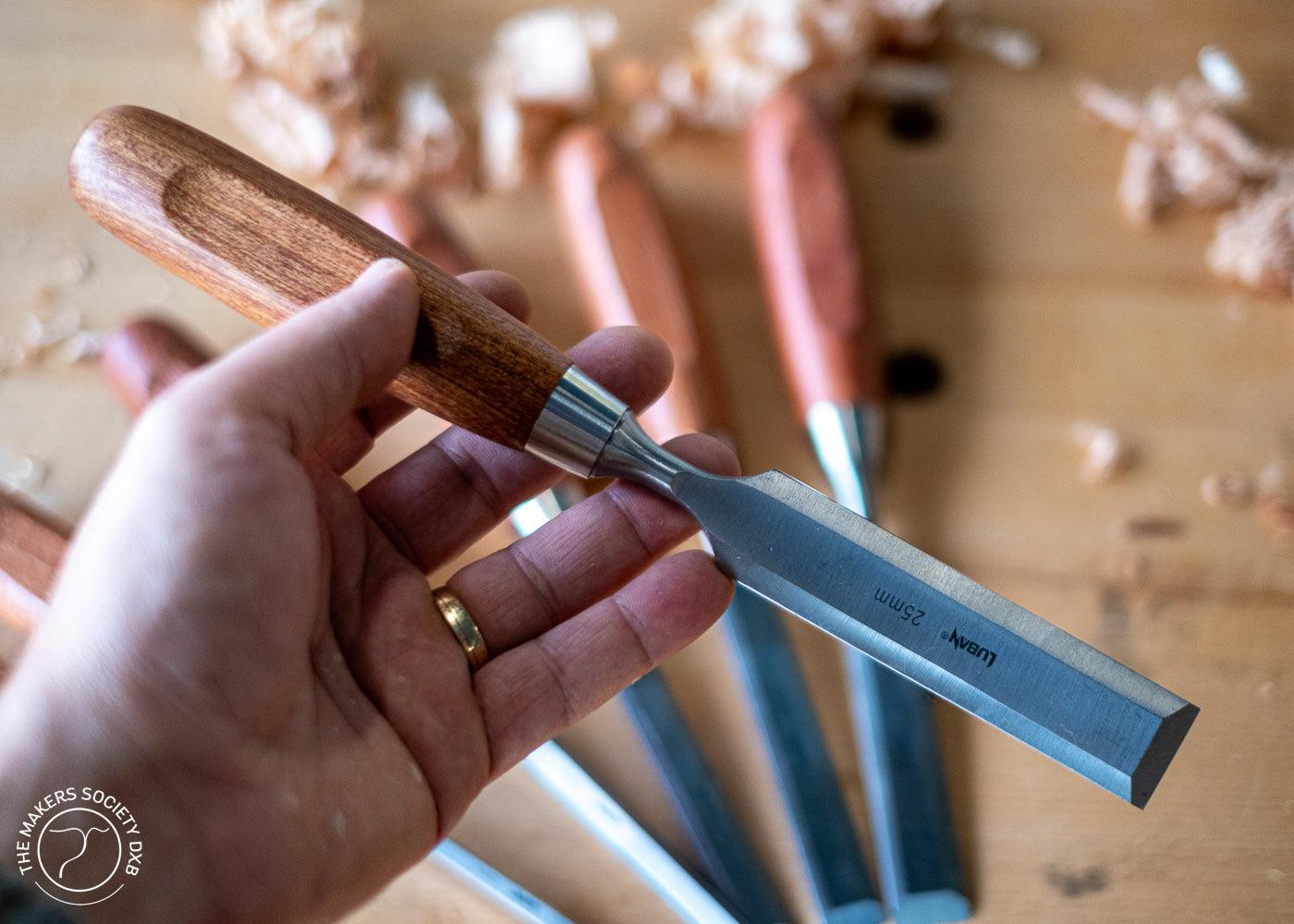 Close-up of a hand holding a Luban bench chisel with wooden handle and sharp metal blade, set against a background of five similar chisels and wood shavings