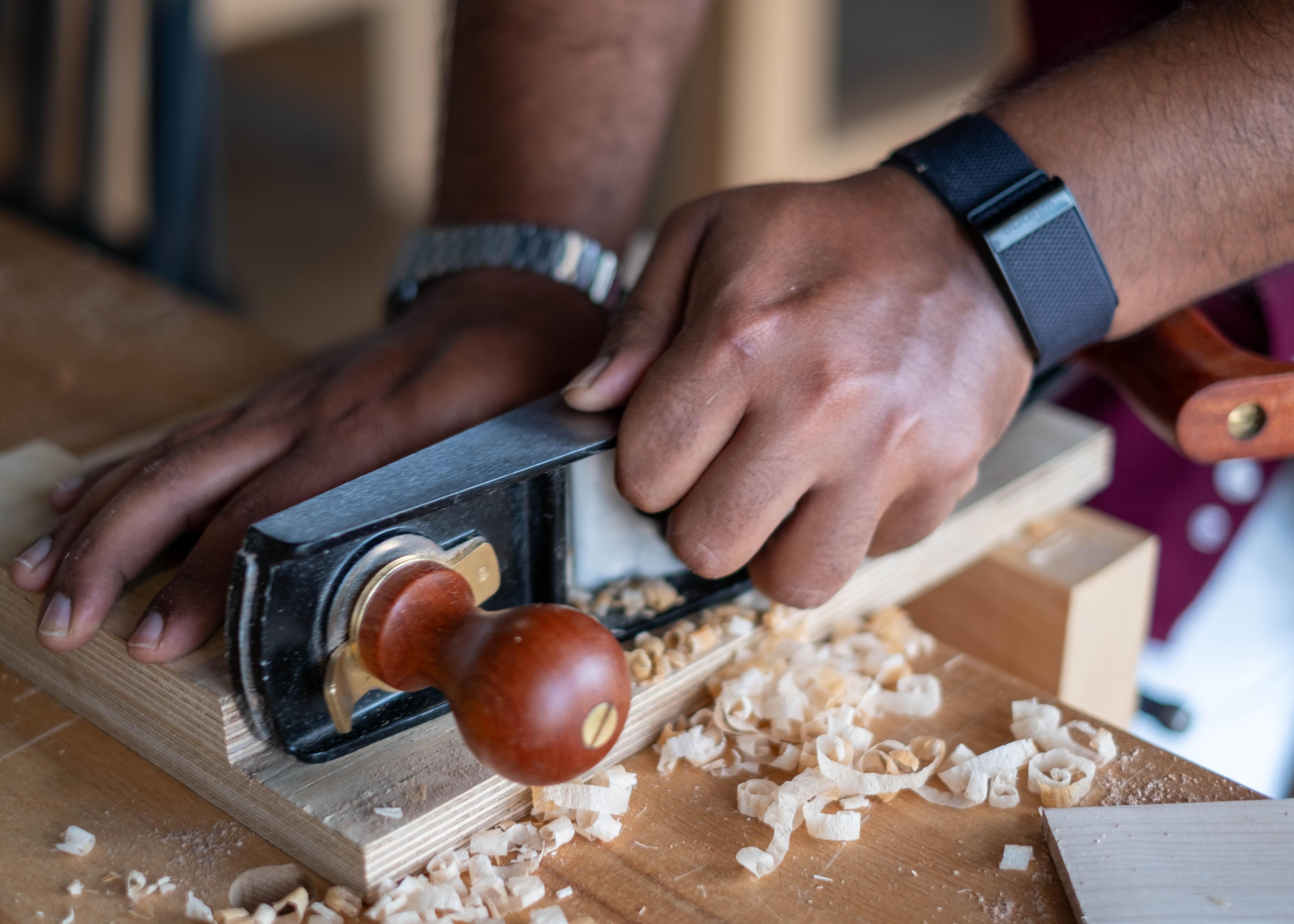 Close-up of hands using a wooden-handled hand plane to shave wood shavings on a woodworking bench