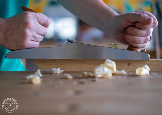 Close-up of hands using a Luban low-angle jack plane to smooth wood with shavings on a workbench
