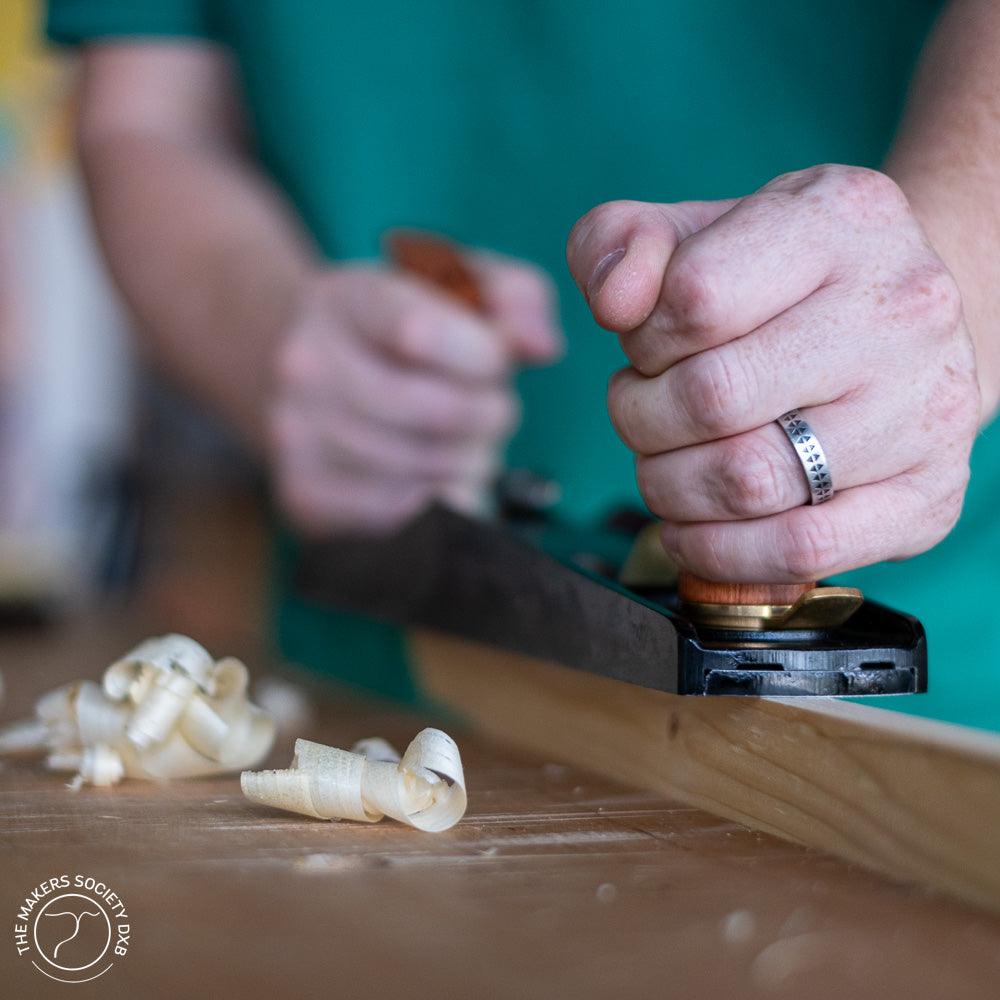 Close-up of hands using a low-angle jack plane on wood, creating curled shavings on a workbench
