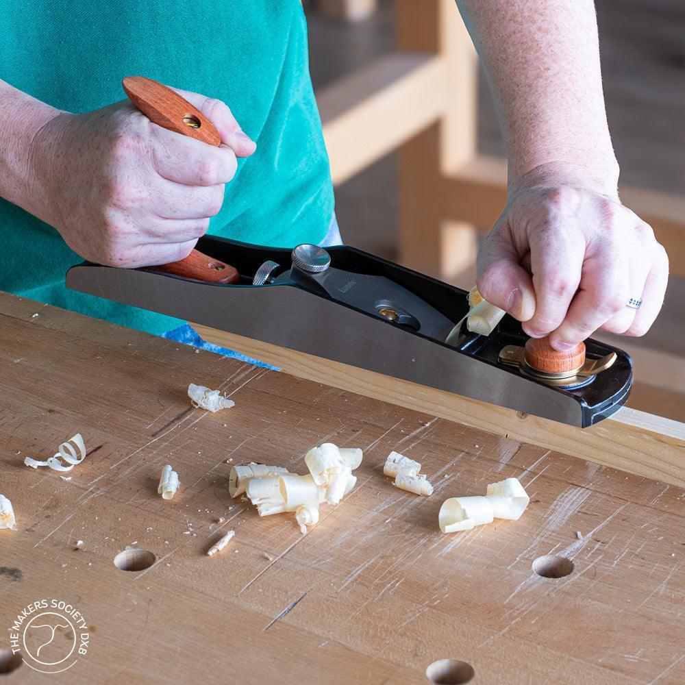 Hands using a Luban 5 low-angle jack plane to shave wood shavings on a workbench
