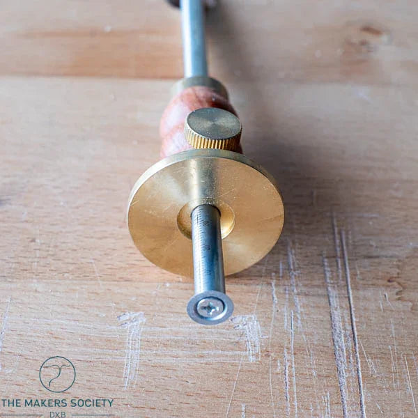 Close-up of a brass and wood marking gauge tool for woodworking on a wooden surface with The Makers Society DXB logo