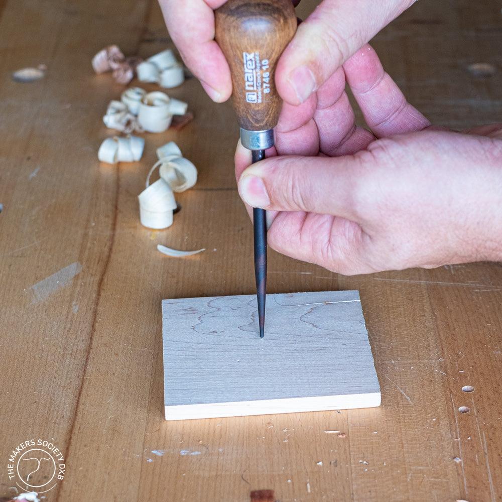 Hand holding Narex conical awl with wooden handle, piercing light wood block on workbench with wood shavings