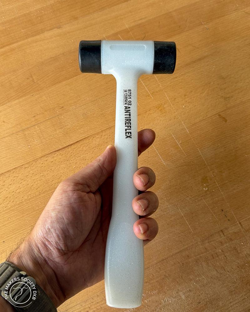 Hand holding a white Narex ANTIRFLEX mallet with black plastic faces on a wooden surface