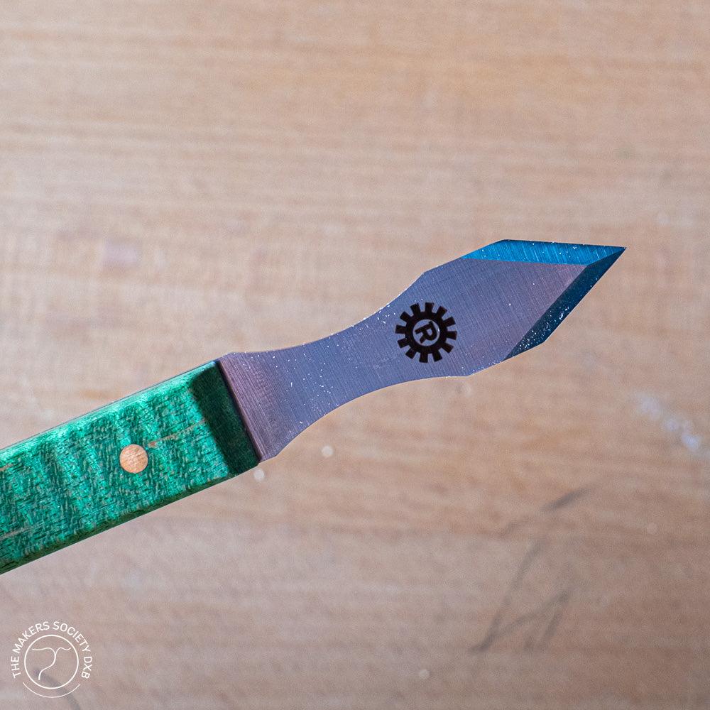 Close-up of a marking knife with a green wooden handle and sharp angled blade, engraved with a gear logo
