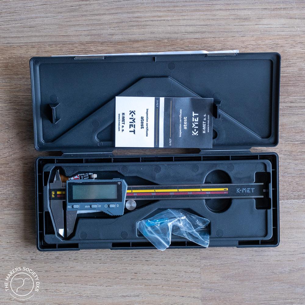 Digital caliper with IP67 rating in open black plastic case, including inspection certificates and accessories on wooden surface