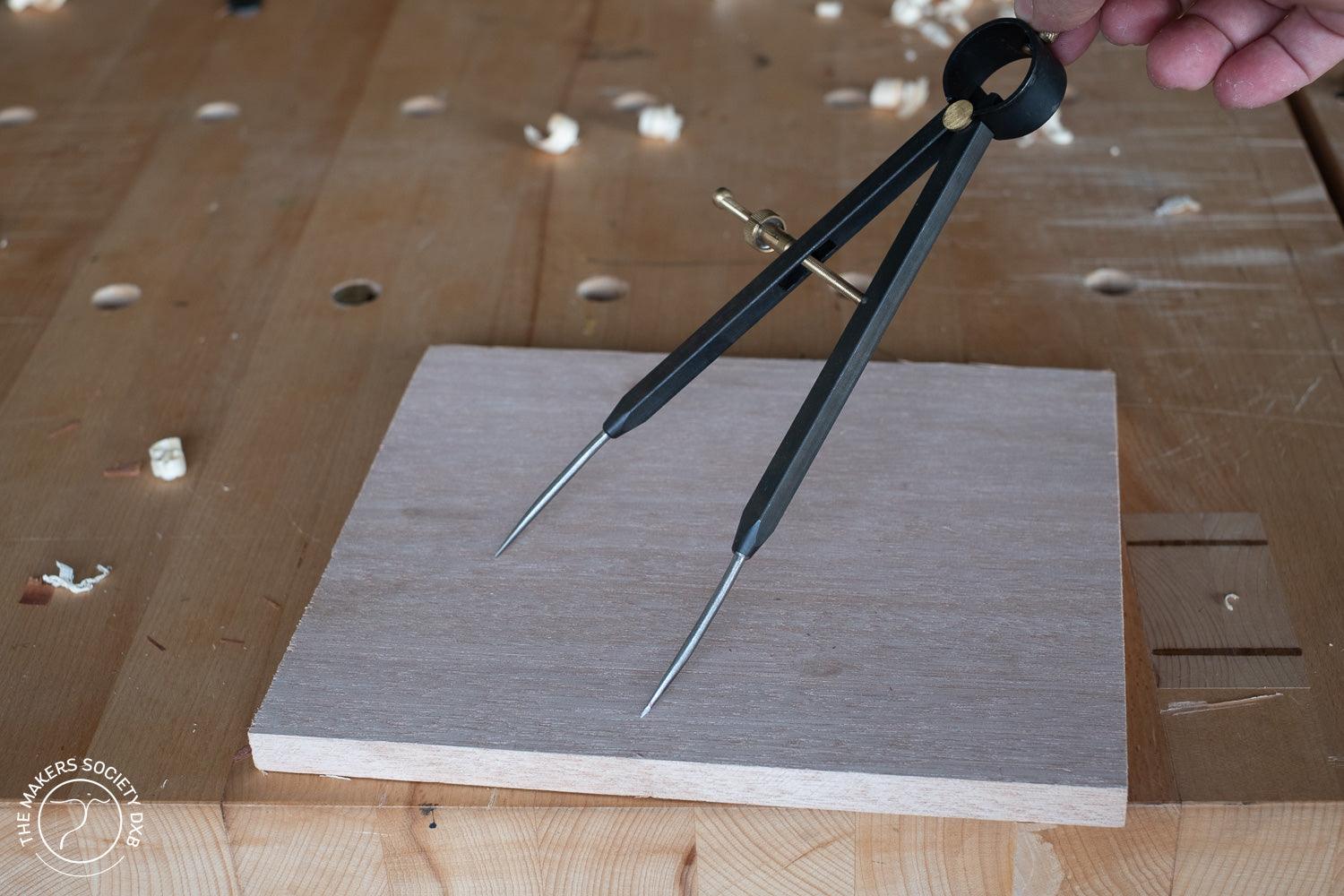 Black spring bow divider tool held over a wooden board on a workbench with wood shavings