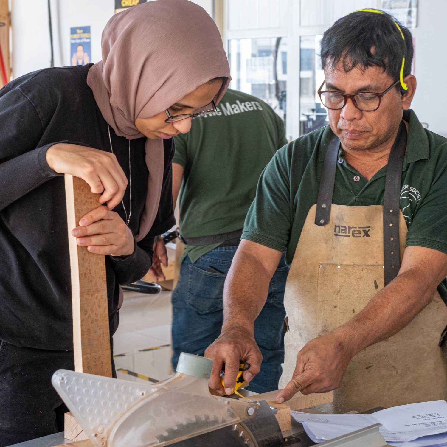 A woodworking tutor points to a specific mark on a piece of timber while explaining the operation of a miter saw to a female student wearing a hijab. Technical drawings and measuring tapes are visible on the workbench as they prepare for a cut.
