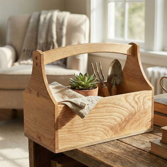 A lifestyle photograph of the handcrafted ash wood toolbox from The Maker's Society, sitting on a rustic wooden table in a sunlit living room. It holds a small potted succulent, a folded linen cloth, and two vintage gardening tools, showcasing its use as home decor.