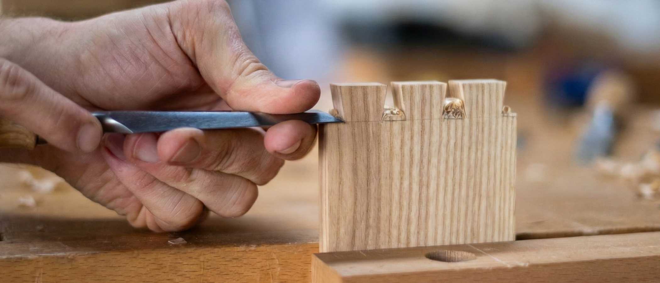 A close-up shot focusing on a woodworker's hands using a sharp metal chisel to refine a dovetail joint. Fine wood shavings are visible on the workbench, highlighting the precision of the hand-tool work.