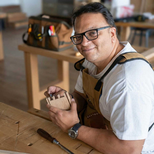 A woodworker at The Makers Society Dubai wearing glasses and a tan work apron smiles at the camera while holding a small, hand-crafted wooden joinery piece at a workbench in a woodworking studio.