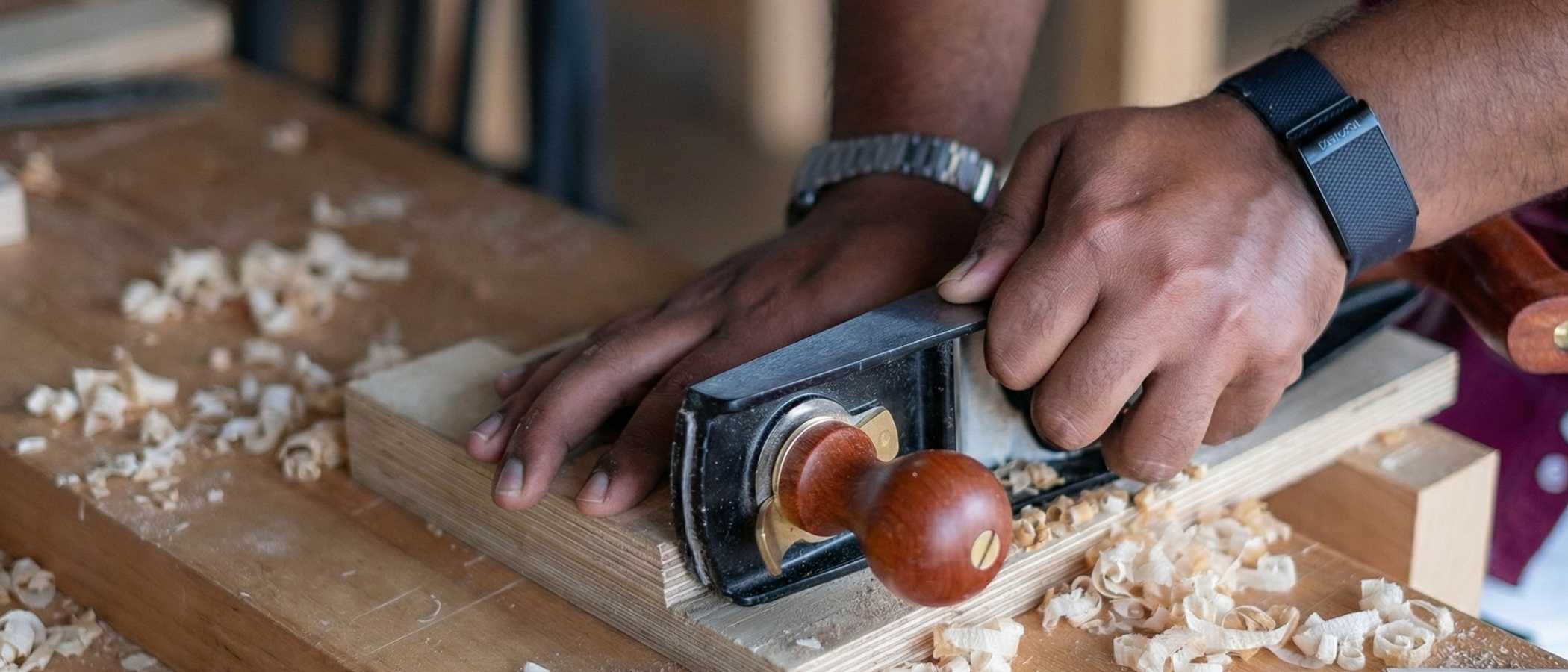 Detailed view of a woodworker’s hands operating a manual hand plane across a wooden board. Fresh, curled wood shavings are scattered across the workbench around the tool.