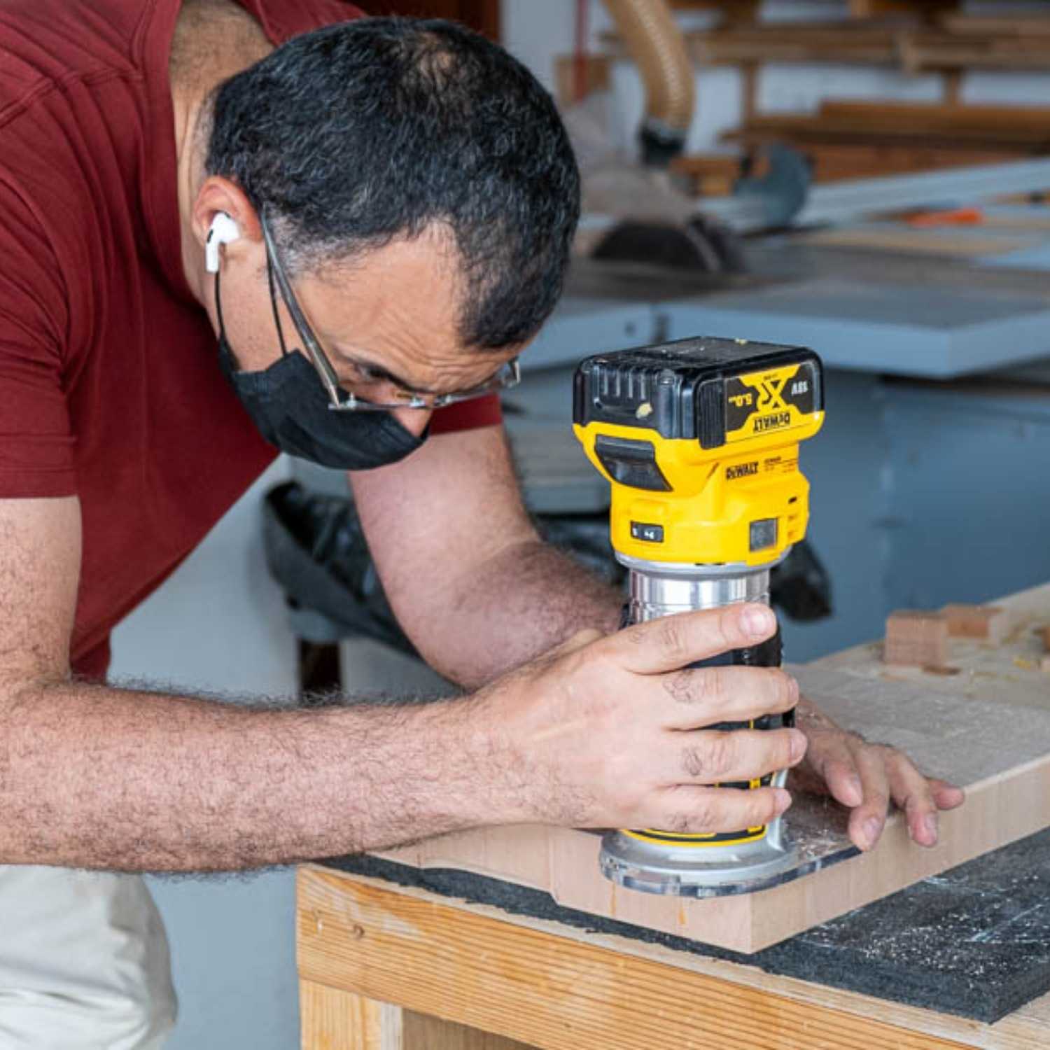 A man wearing a maroon shirt and a black face mask operates a yellow DeWalt compact power router. He is carefully guiding the tool along the edge of a thick wooden board to create a profile, with wood shavings visible on the workbench.