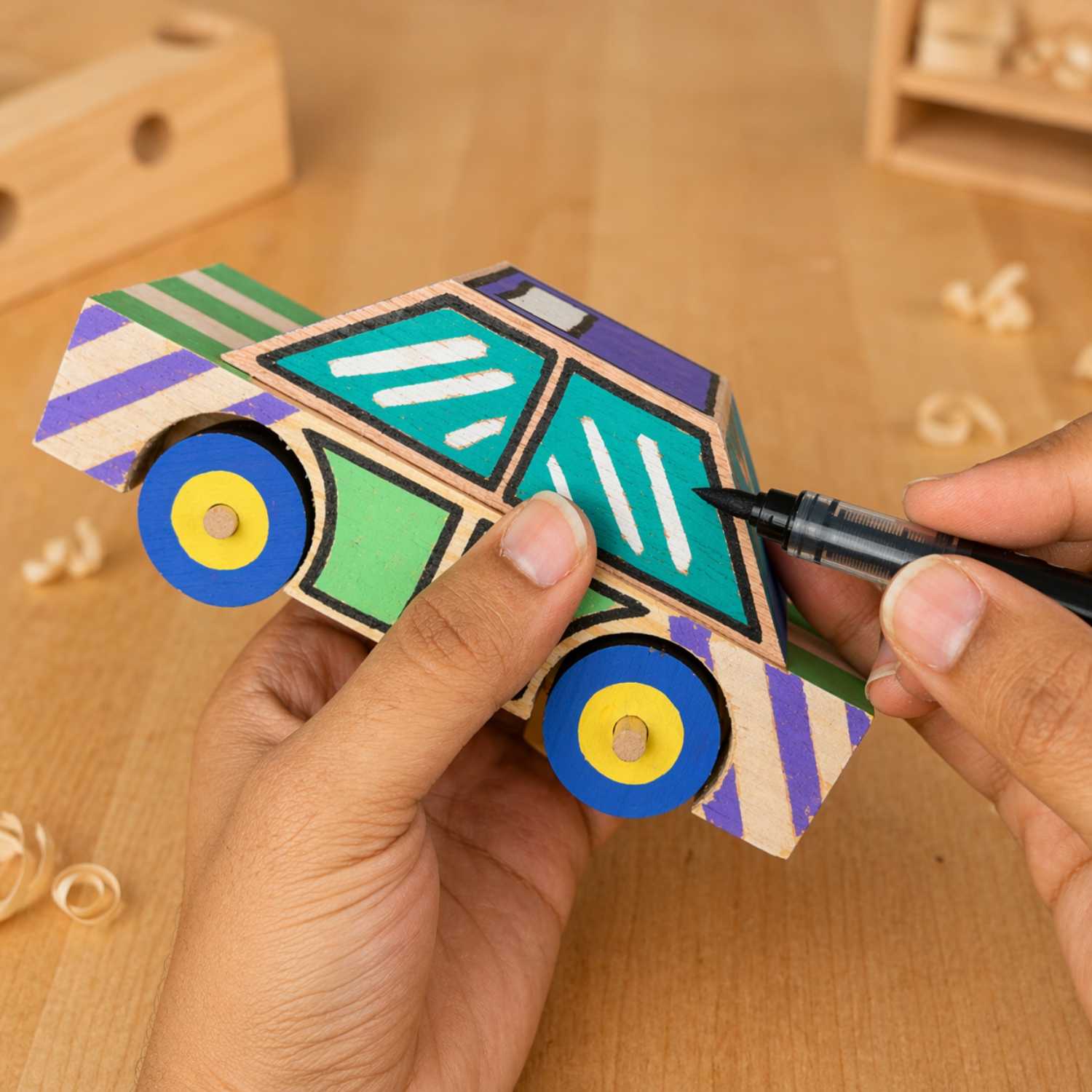 Close-up of hands decorating The Makers Society wooden taxi car kit with a black marker on a workshop table.