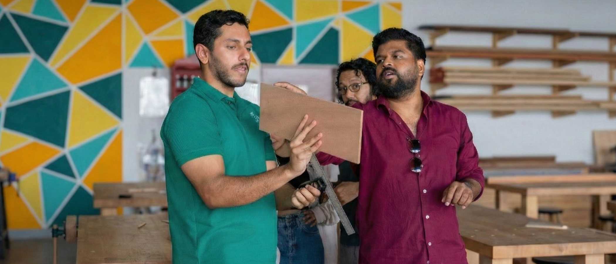 Tutor and student at The Makers Society Dubai discuss a project. The tutor in a green shirt holds a piece of wood while the student uses a combination square to check measurements. A vibrant yellow and teal geometric mural is visible in the background.